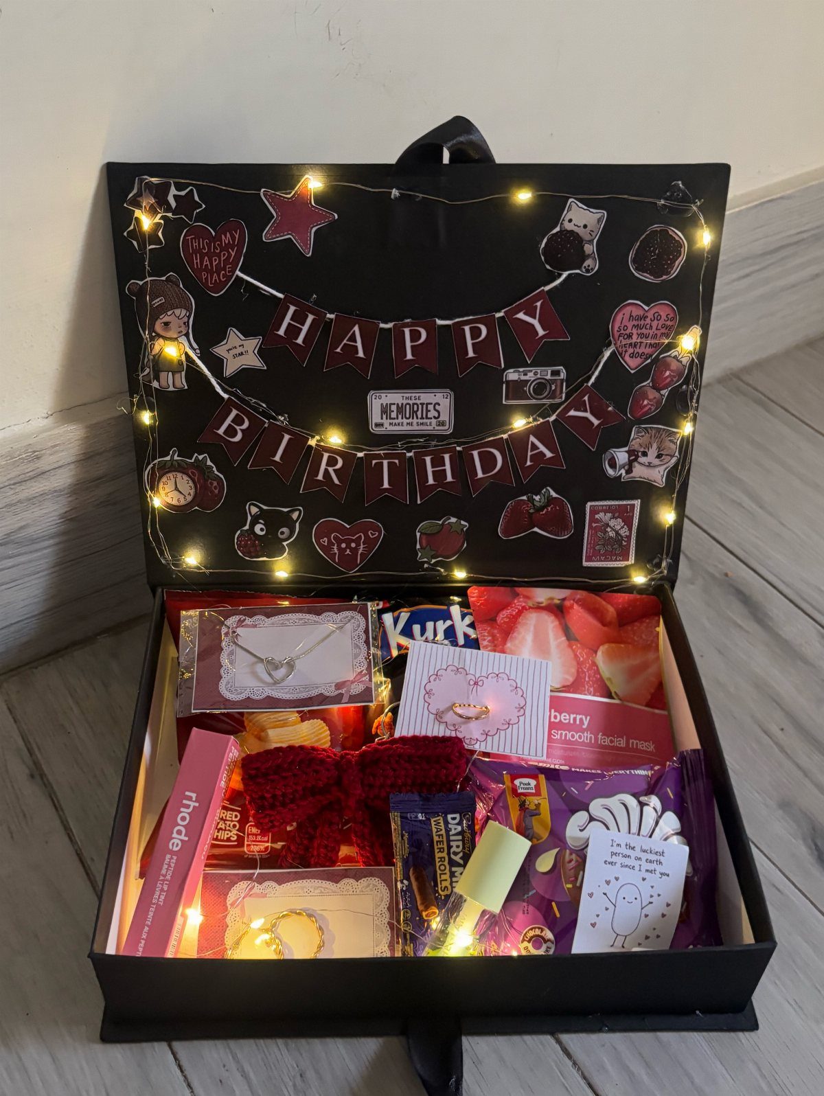 Open black birthday gift box filled with treats and decorated with fairy lights.