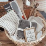 Woven gift basket containing kitchen towels, wooden utensils, and a decorative framed sign.
