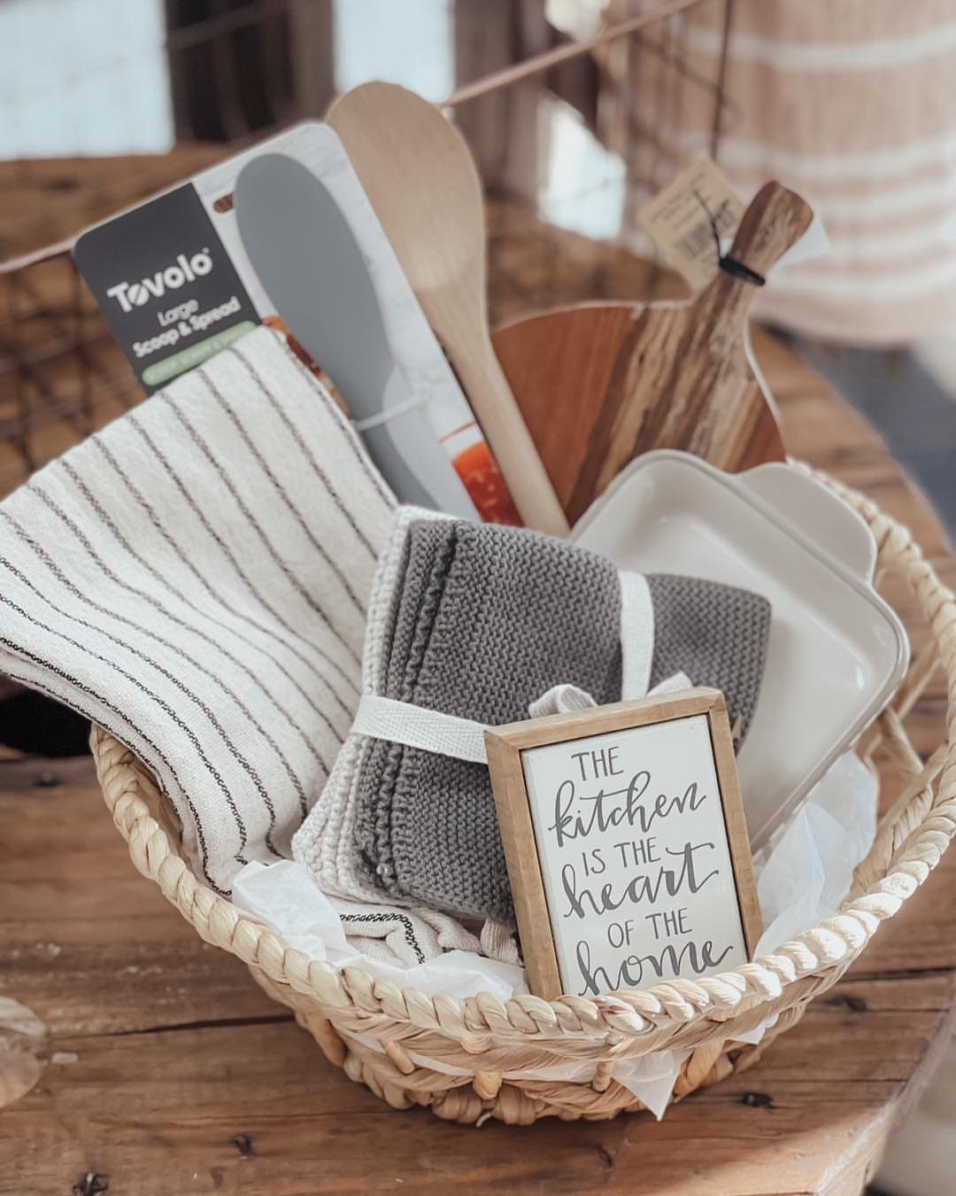 Woven gift basket containing kitchen towels, wooden utensils, and a decorative framed sign.