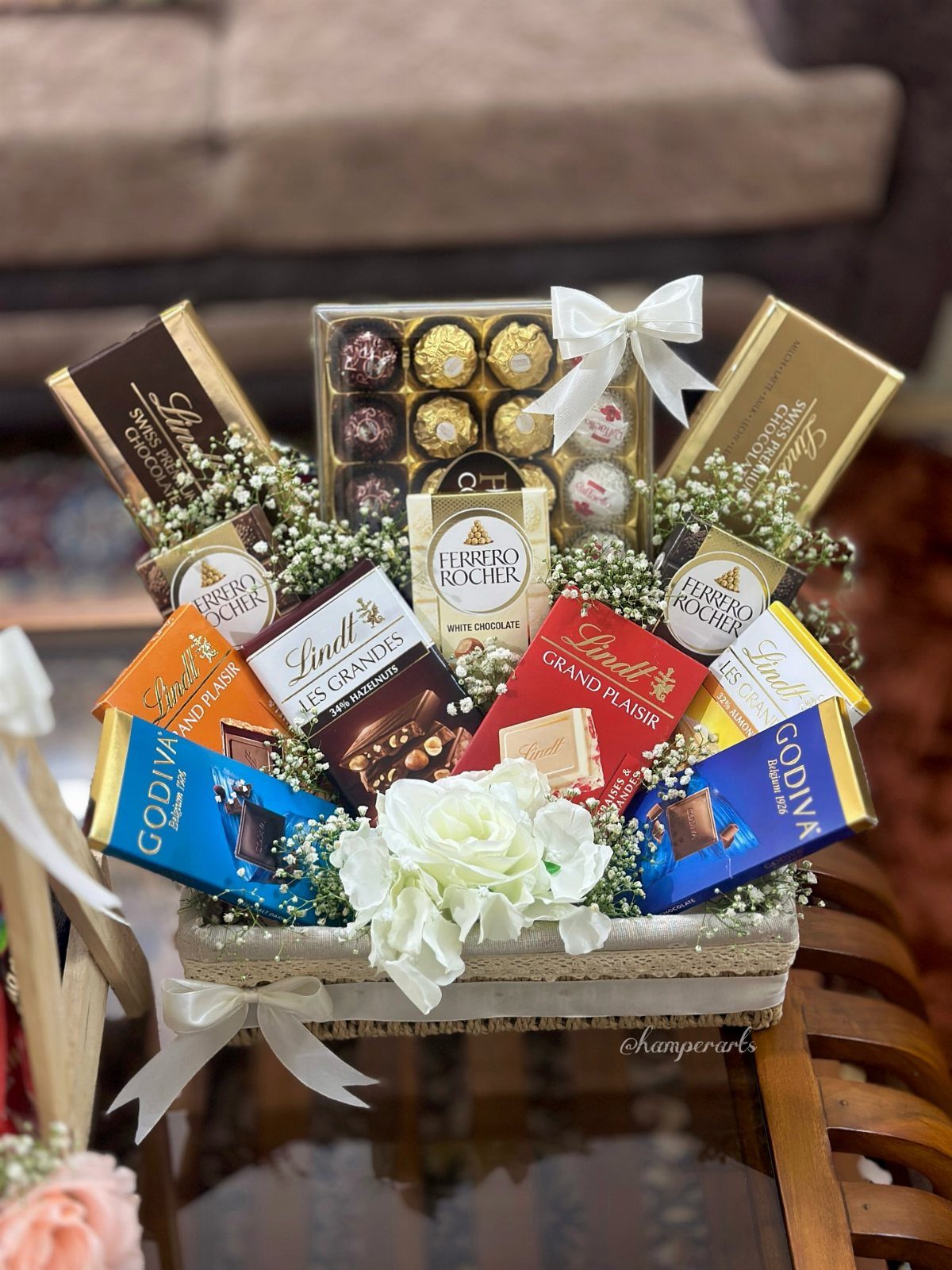 Woven gift basket filled with assorted chocolate bars and truffles decorated with white flowers.