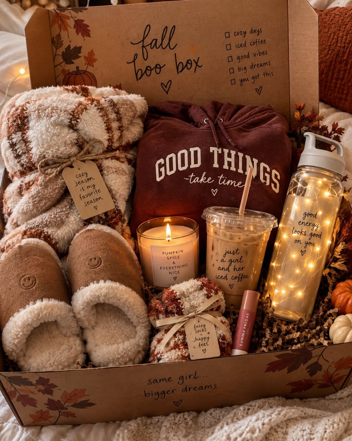 Gift box containing a plaid blanket, maroon hoodie, fluffy slippers, and autumn-themed accessories.