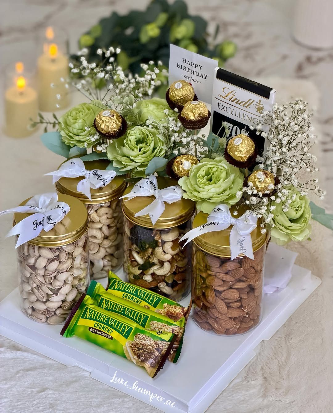 Gourmet food gift hamper with nut jars, chocolates, granola bars, and green flowers.