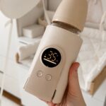 Beige portable baby bottle warmer with digital display held in a hand.