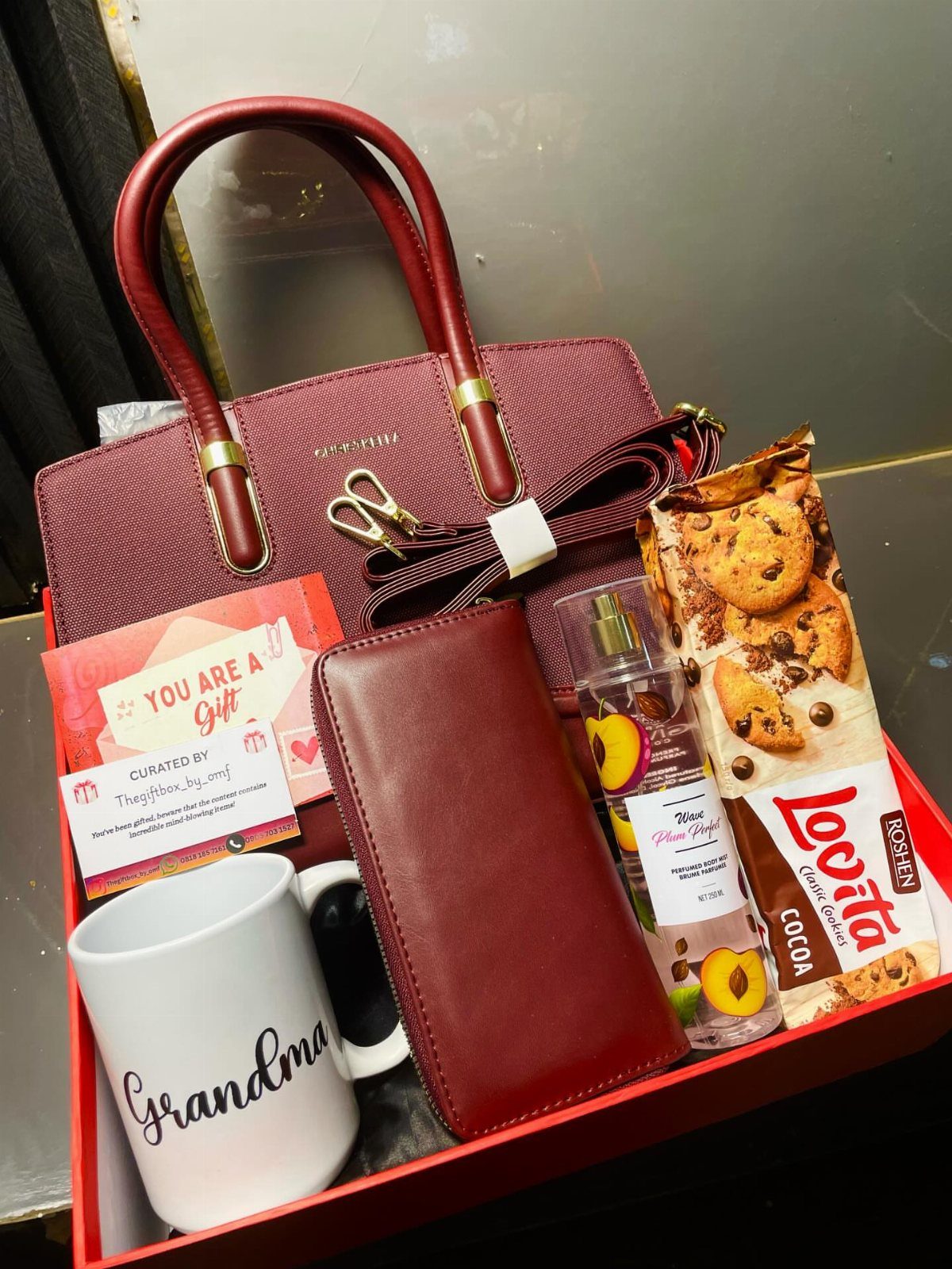 Gift box for grandma with burgundy handbag, wallet, white mug, and cookies.