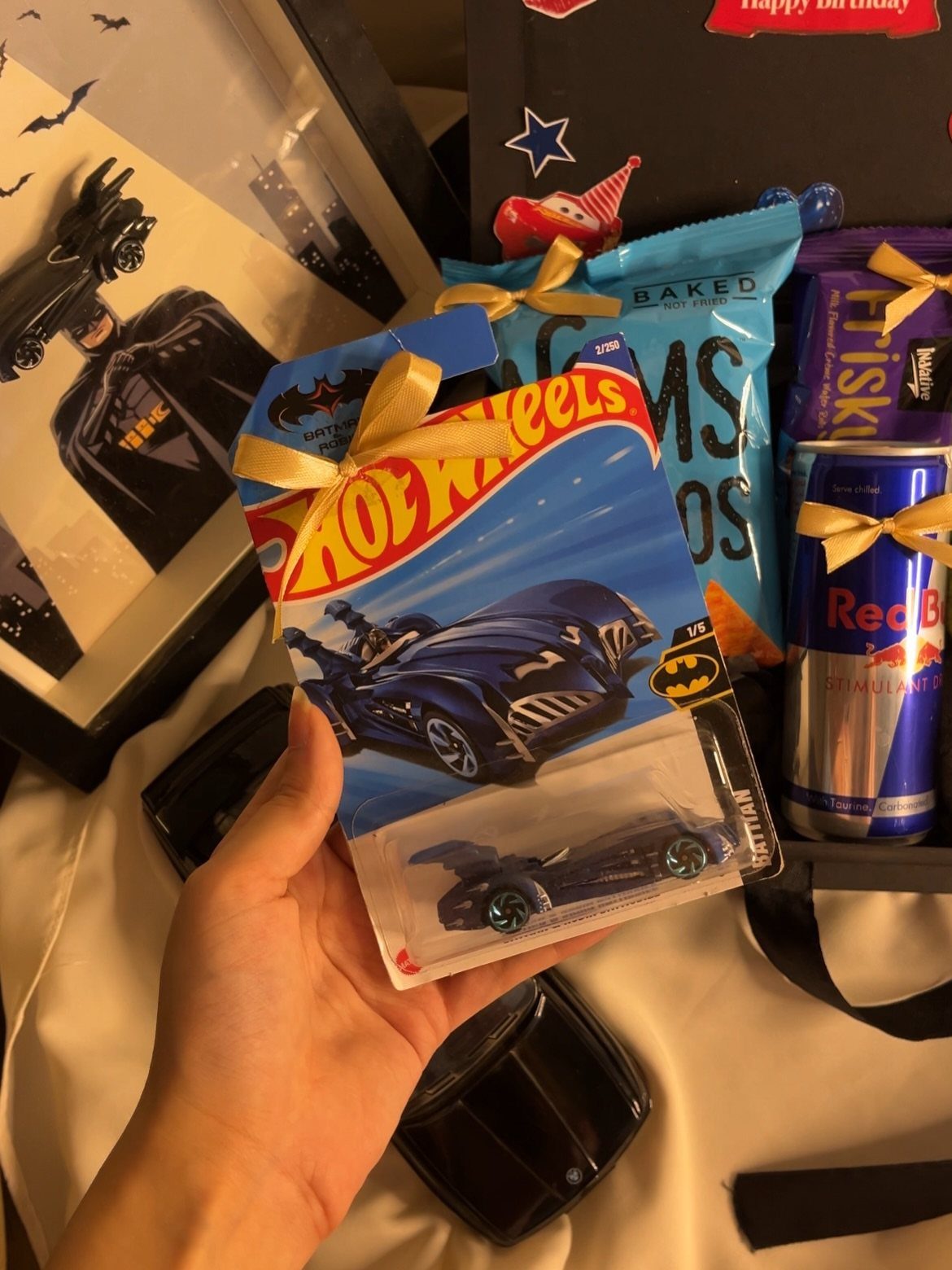 Hand holding a Hot Wheels Batman car among birthday gifts and snacks.