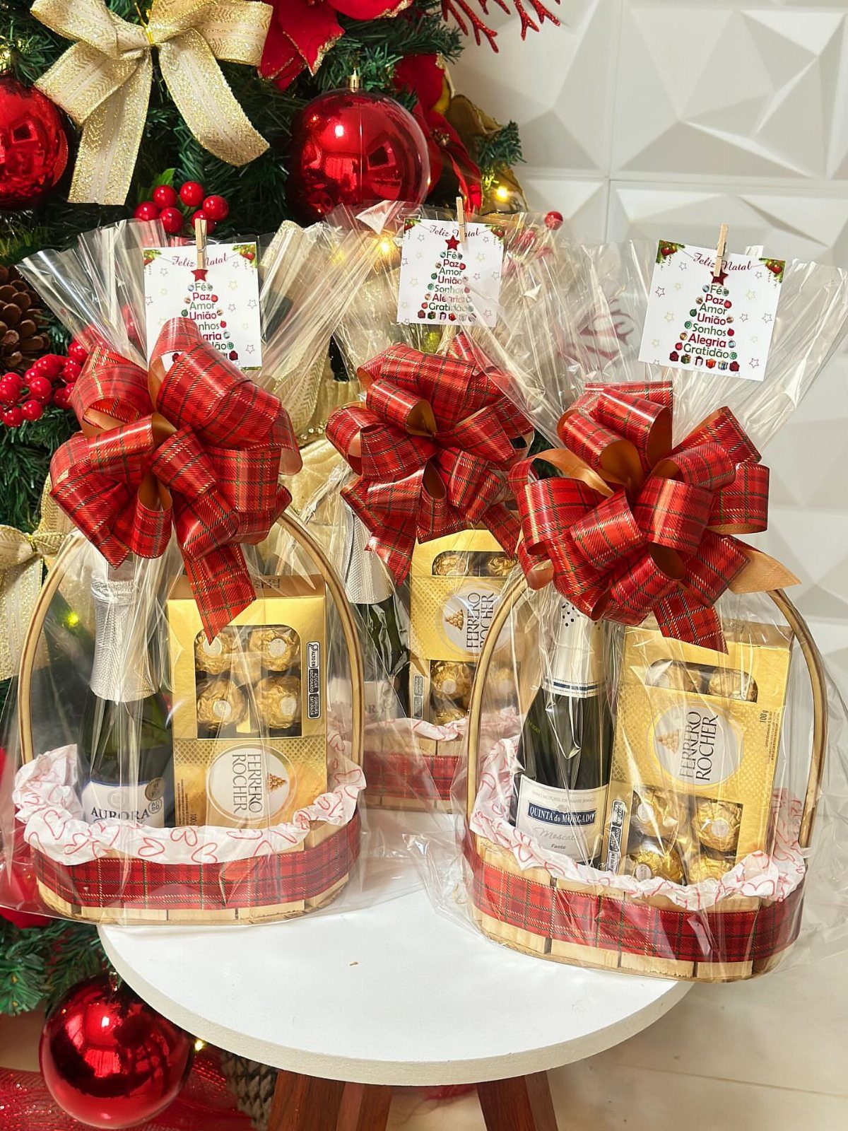 Three Christmas wine gift baskets with red bows and chocolates by a tree