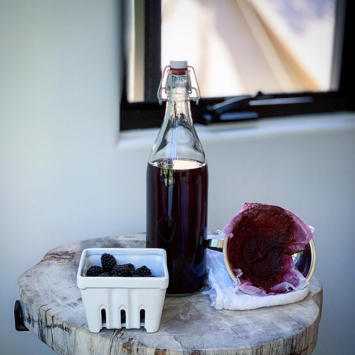 Dark purple homemade liqueur in a glass bottle with fresh blackberries and strainer.