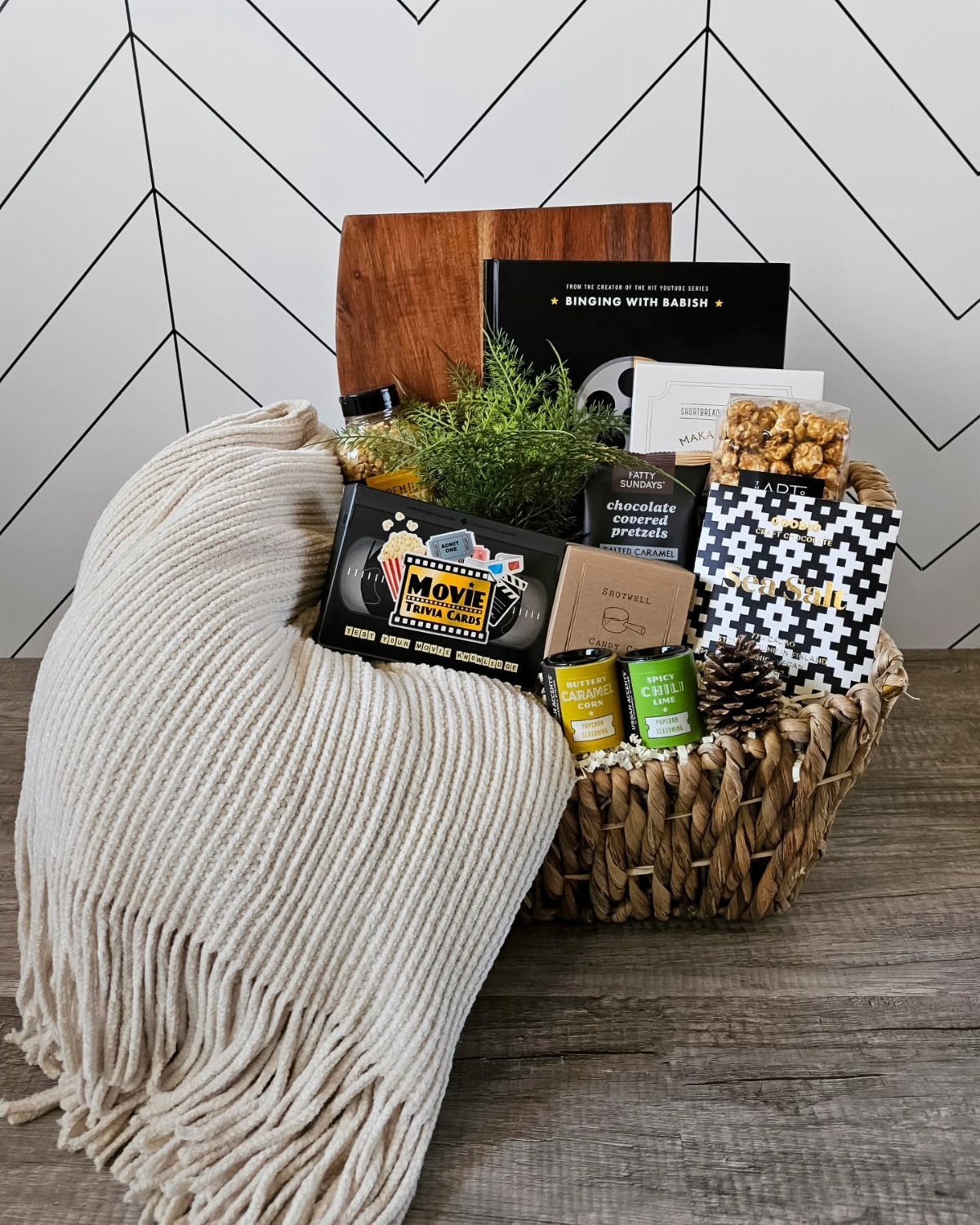 Woven gift basket with popcorn, movie snacks, a book, and a cream throw blanket.