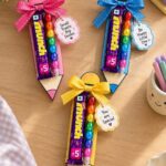 Pencil-shaped gift holders containing chocolate bars and colorful markers with decorative ribbons.