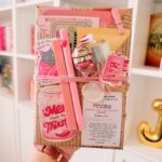 Brown paper gift bag filled with pink stationery, candy, and book-themed accessories.