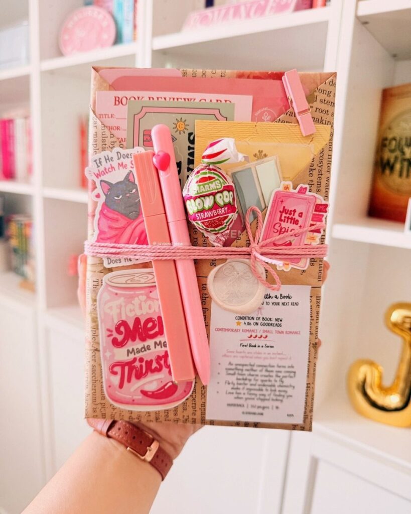 Brown paper gift bag filled with pink stationery, candy, and book-themed accessories.